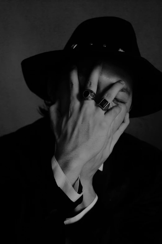 a black and white photo of a man in a hat, a black and white photo, tumblr, his hands buried in his face, rings, album cover, faceless people dark