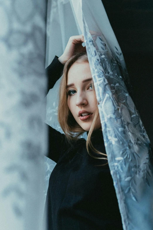 a woman standing behind a curtain in front of a window, inspired by Elsa Bleda, pexels contest winner, beautiful taissa farmiga, wrapped in a black scarf, blue and gray colors, sydney sweeney