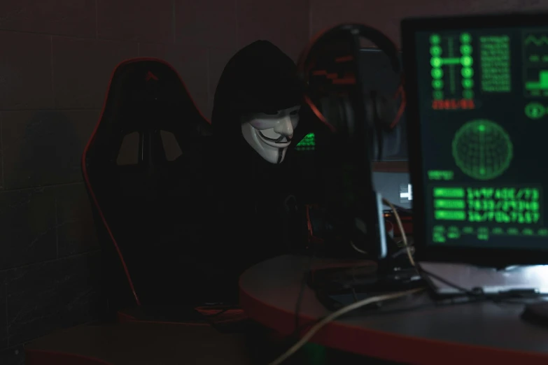 a person wearing a mask sitting in front of a computer, cryptic, 15081959 21121991 01012000 4k, black hood, high quality photo