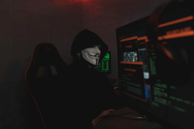 a person wearing a mask sitting in front of a computer, by Adam Marczyński, pexels contest winner, serial art, black hood, crypto, halloween, with screens and silks