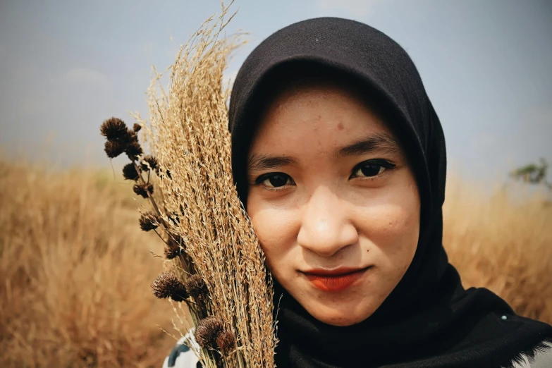 a woman standing in a field holding a bunch of dried plants, pexels contest winner, hurufiyya, south east asian with round face, avatar image, islamic, headshot profile picture