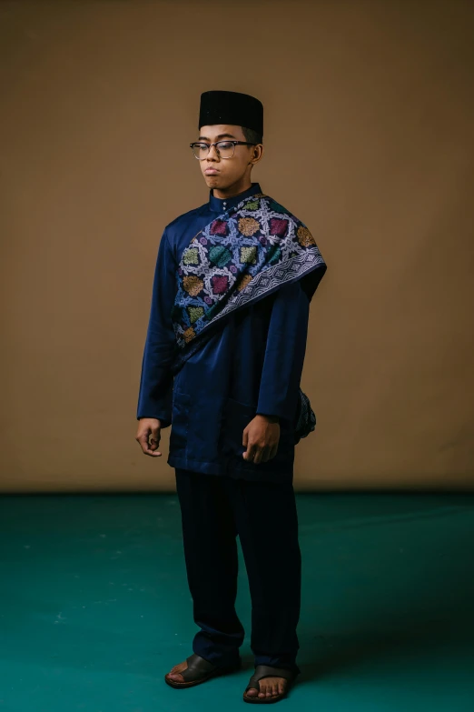 a man standing in front of a brown wall, an album cover, by Basuki Abdullah, gold brocaded dark blue clothes, non binary model, wearing an academic gown, portrait of a patchwork boy