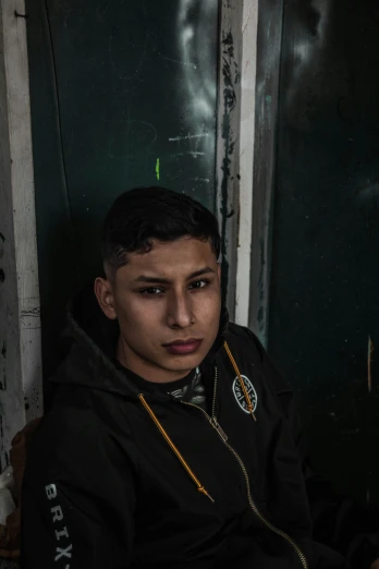 a young man sitting in front of a green door, eloy morales, 1 7 - year - old boy thin face, dark backdrop, eytan zana