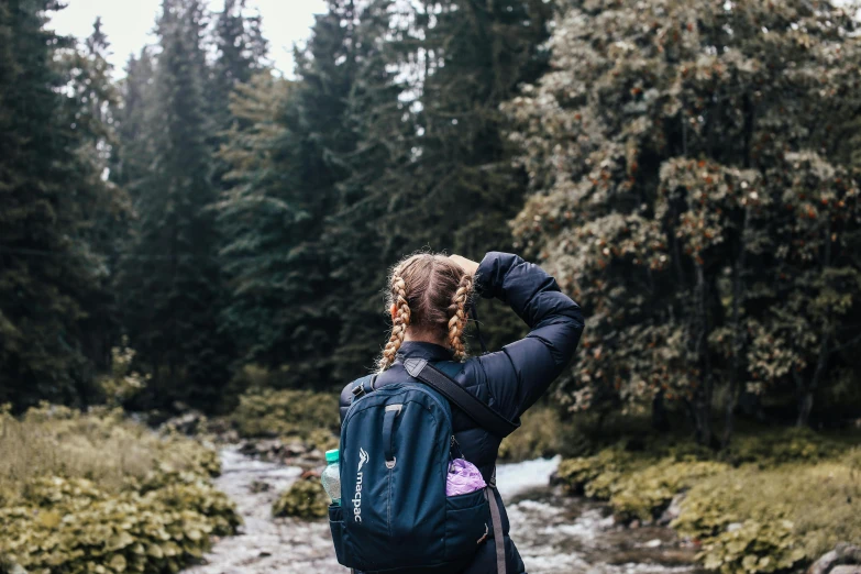 a woman with a backpack standing in front of a stream, by Emma Andijewska, pexels contest winner, pine trees in the background, waving, greta thunberg, seen from the back