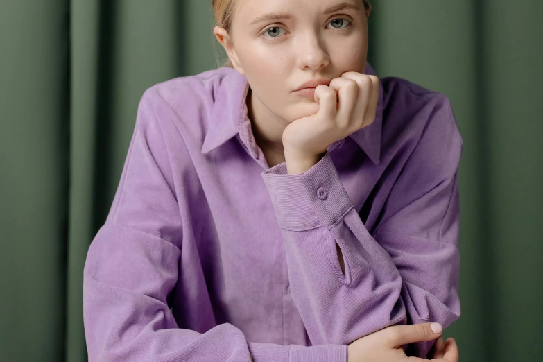 a woman sitting at a table with her hand on her chin, inspired by Violet Fuller, trending on pexels, long shirt, nina tryggvadottir, purple clothes, teenager girl