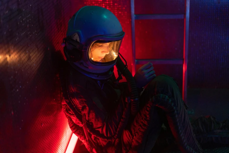 a man in a helmet sitting against a wall, inspired by roger deakins, girl in space, red shift, 8 0 s asian neon movie still, natalia dyer