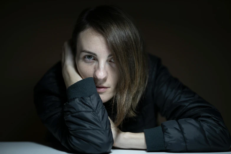 a woman sitting at a table in the dark, a portrait, inspired by Elsa Bleda, pexels contest winner, hyperrealism, model wears a puffer jacket, bored expression, girl with brown hair, 30 years old woman