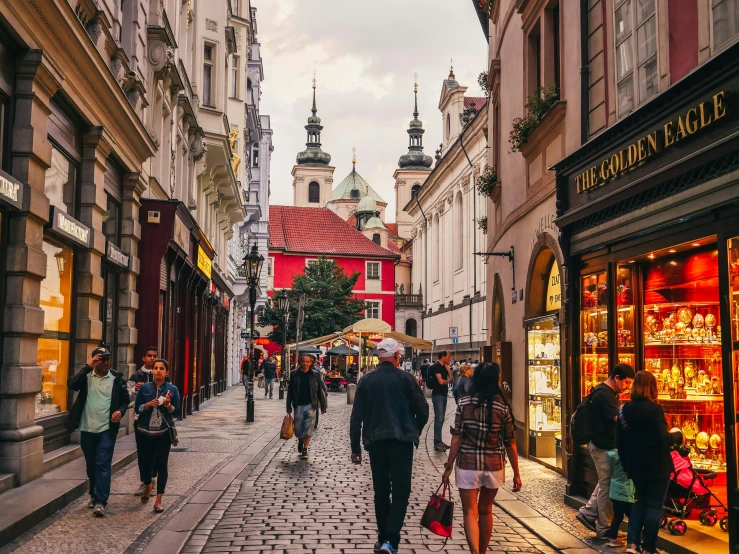 a group of people walking down a street next to tall buildings, by Emma Andijewska, pexels contest winner, art nouveau, baroque winding cobbled streets, slovakia, olive green and venetian red, square