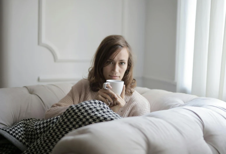 a woman sitting on a couch holding a coffee cup, by Alice Mason, pexels, happening, freezing, white, worried, grey