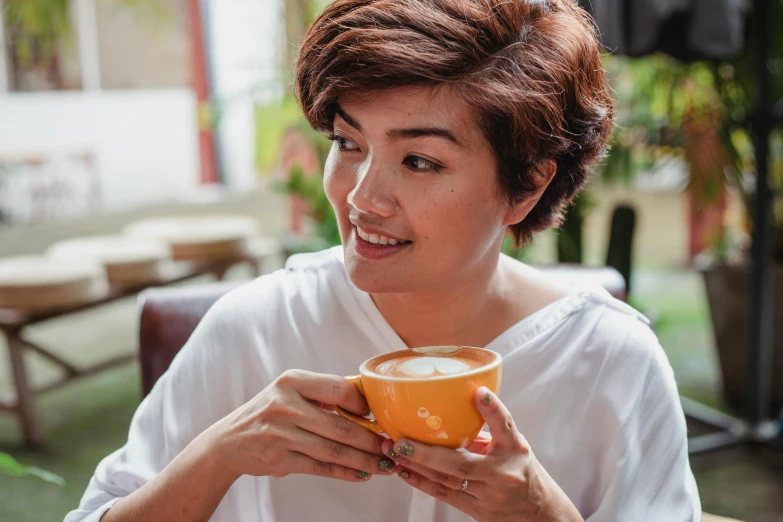 a woman sitting at a table with a cup of coffee, inspired by Ruth Jên, happening, wavy short hazel hair, vietnamese woman, orange hue, profile image