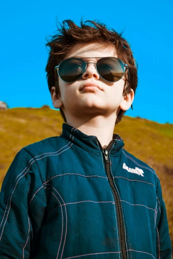 a young boy wearing sunglasses standing in front of a hill, model wears a puffer jacket, zoomed in, avatar image, performance