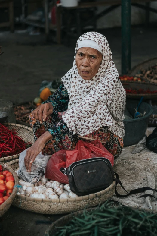 a woman sitting in front of baskets of vegetables, a picture, by Matija Jama, pexels contest winner, sumatraism, wearing a head scarf, old lady cyborg merchant, bags on ground, gif