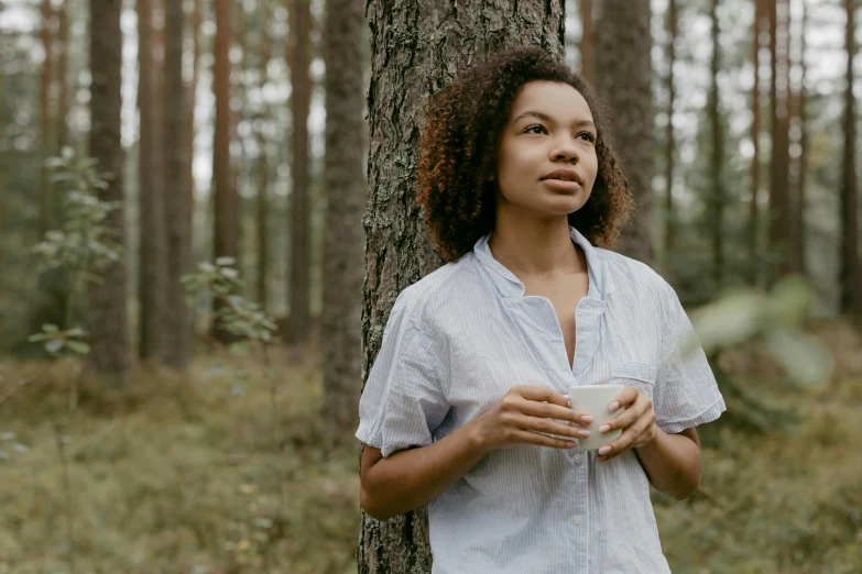 a woman standing next to a tree holding a cup, by Adam Marczyński, pexels contest winner, mixed-race woman, wearing a linen shirt, avatar image, forested