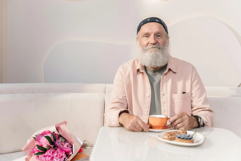 a man sitting at a table with a cup of coffee, overalls and a white beard, with flowers, avatar image, while smiling for a photograph