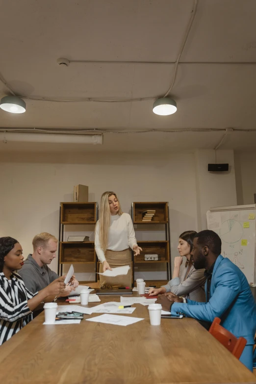 a group of people sitting around a wooden table, in an office