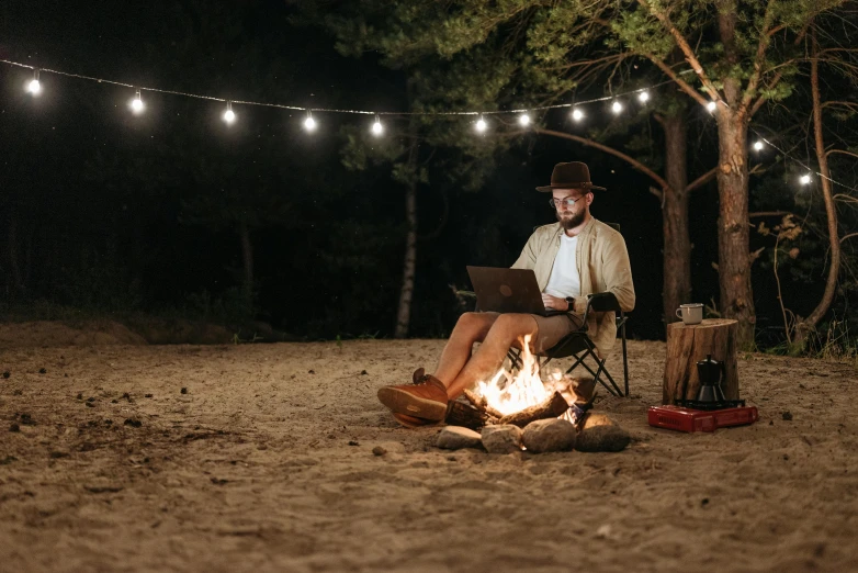 a man sitting in a chair next to a campfire, trending on pexels, sitting at a computer, string lights, aboriginal australian hipster, hiking clothes