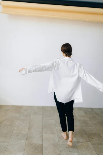 a woman in a white shirt standing in a room, inspired by Josefina Tanganelli Plana, unsplash, light and space, arms stretched out, in front of white back drop, walking to the right, cropped wide sleeve