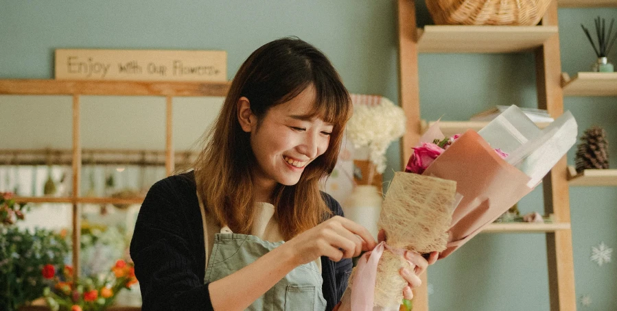 a woman in an apron holding a bunch of flowers, pexels contest winner, asian woman made from origami, smiling playfully, flower shop scene, ribbon