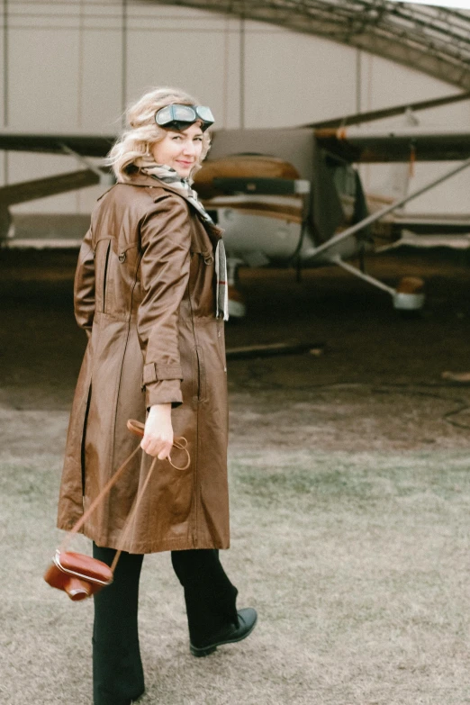 a woman standing in front of an airplane, inspired by Harrison Fisher, pexels contest winner, light brown trenchcoat, costume, brown, field