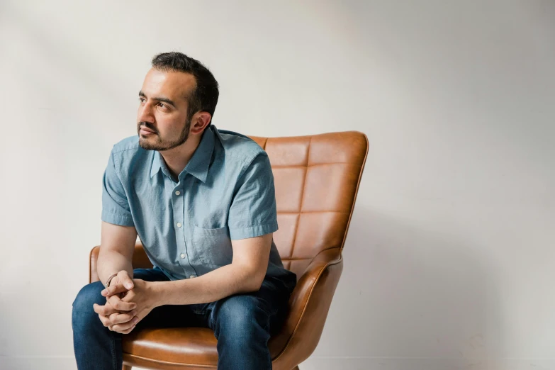 a man sitting on top of a brown chair, a portrait, inspired by Alejandro Obregón, pexels contest winner, wearing a shirt and a jean, clear julian lage face, looking to the side off camera, architect