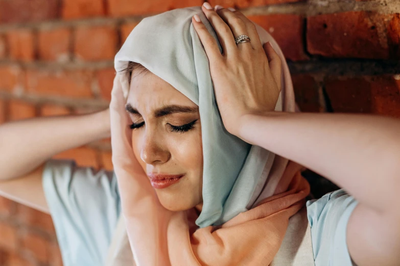 a woman wearing a hijab in front of a brick wall, trending on pexels, hurufiyya, stressed expression, pastel hues, wearing a head scarf, hands in her hair