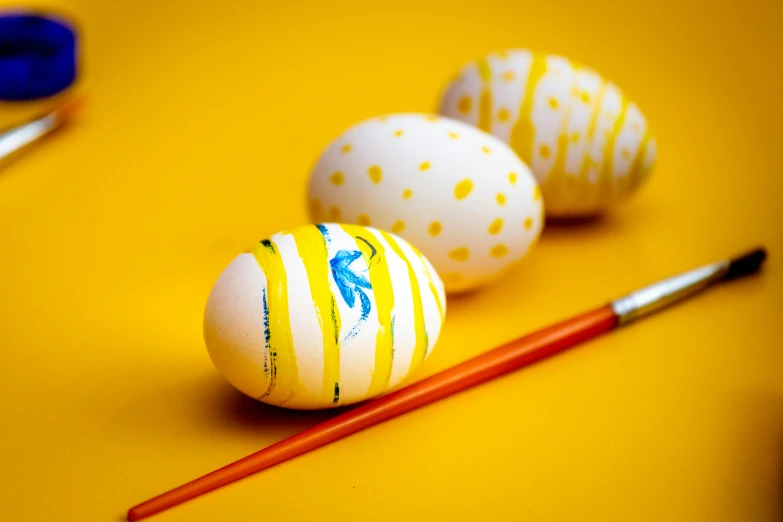 three painted eggs and a paintbrush on a yellow surface, an album cover, by Maksimilijan Vanka, pexels, avatar image, cross - hatching, decorated ornaments, 3 4 5 3 1