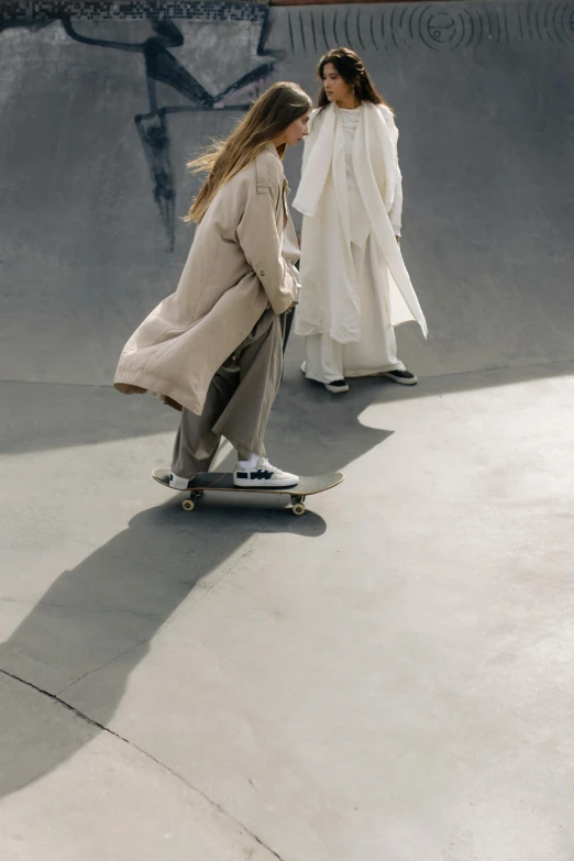 two women riding skateboards at a skate park, trending on unsplash, hyperrealism, wearing long white robe, issey miyake, wearing a long beige trench coat, featured on vogue