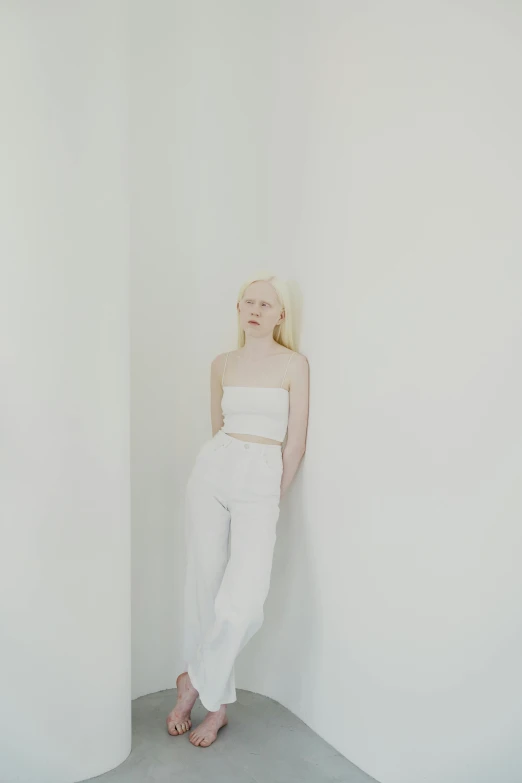 a woman leaning against a wall in a white room, inspired by Vanessa Beecroft, unsplash, albino dwarf, wearing a cropped top, white pants, elle fanning)