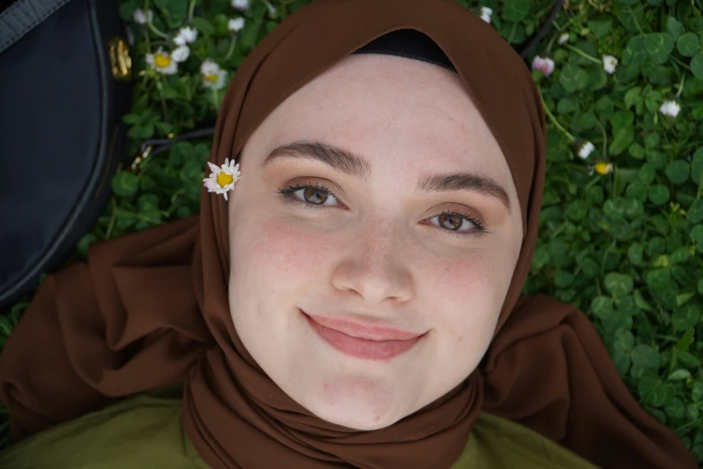 a woman laying in the grass with a flower in her hair, inspired by Nazmi Ziya Güran, hurufiyya, wearing a brown, closeup headshot, avatar image, hijab