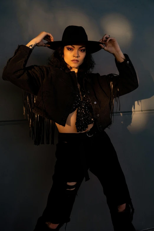 a woman standing in front of a wall wearing a hat, an album cover, by Robbie Trevino, trending on pexels, renaissance, black jacket | shiny, female cowgirl, shot at night with studio lights, doing a sassy pose