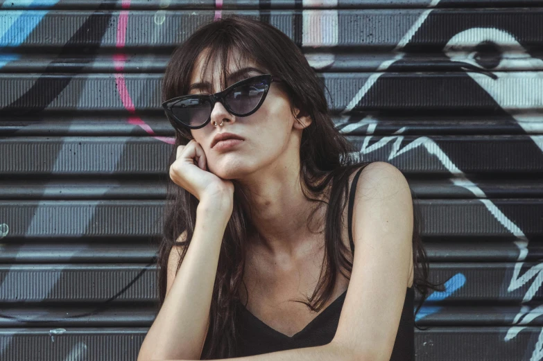 a woman sitting in front of a graffiti covered wall, inspired by Elsa Bleda, unsplash contest winner, black sunglasses, portrait emily ratajkowski, black cat, small square glasses