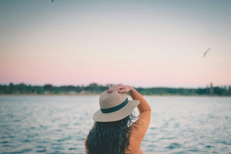 a woman wearing a hat standing in front of a body of water, pexels contest winner, pastel orange sunset, afternoon hangout, over the shoulder, low colour