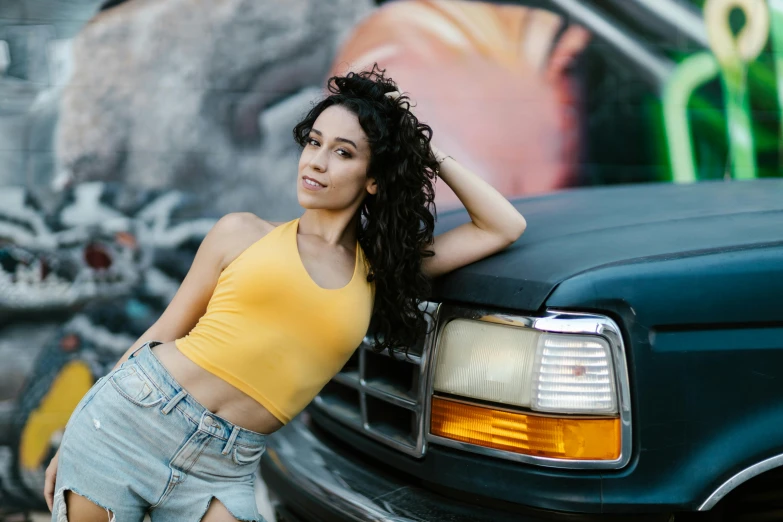 a woman leaning on the hood of a truck, trending on pexels, wearing yellow croptop, curly haired, avatar image, alanis guillen