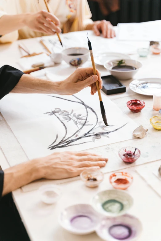 a group of people sitting around a table painting, inspired by Sōami, trending on pexels, process art, chinese ink brush, on a white table, lacquerware, banner