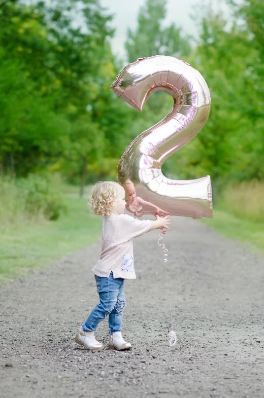 a little girl holding a balloon shaped like the number 2, by David Simpson, happening, rose gold, outdoors, birthday, large tail