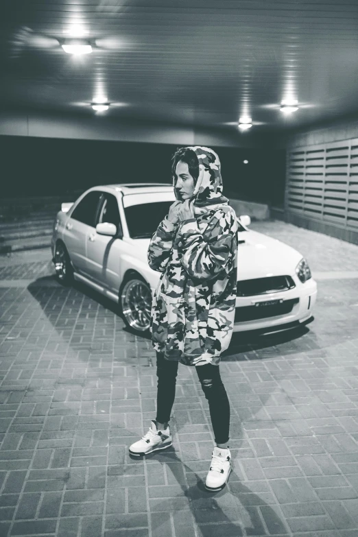 a woman standing in a parking garage next to a car, a black and white photo, by Ismail Acar, unsplash, graffiti, wearing an oversized hoodie, wearing camo, ☁🌪🌙👩🏾, anime car wrap