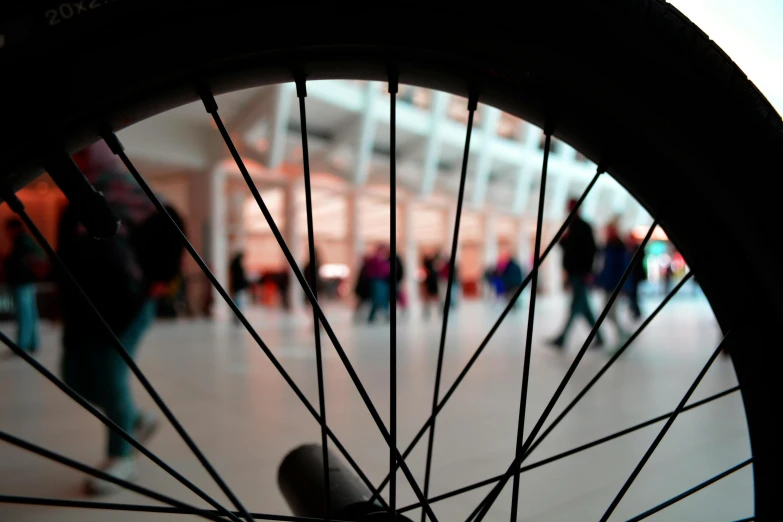a close up of a bicycle wheel with people walking in the background, inspired by Ai Weiwei, pexels contest winner, visual art, brooklyn museum, indoor picture, silhouettes of people, at a mall