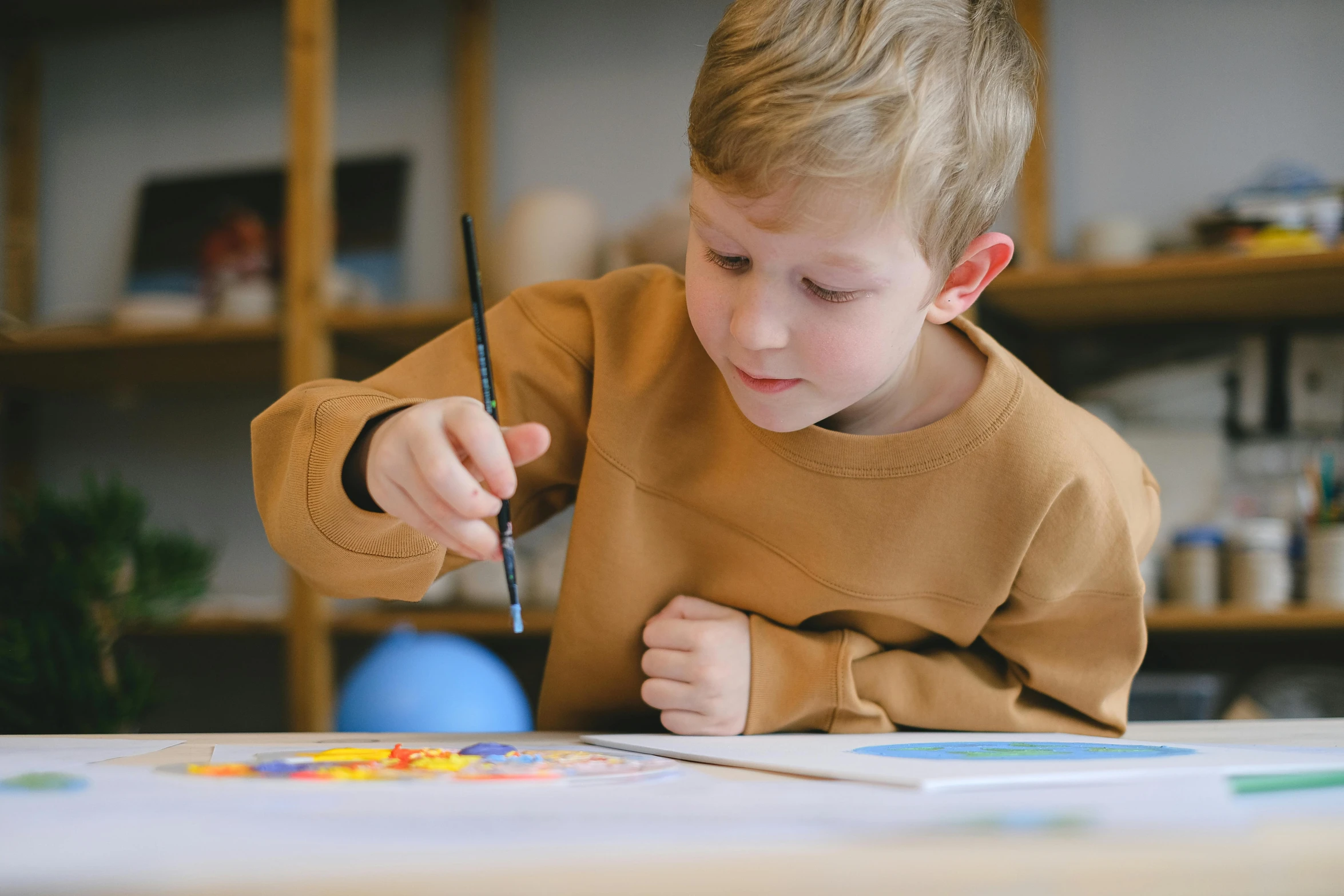 a little boy that is sitting at a table, a child's drawing, inspired by Kyffin Williams, pexels contest winner, visual art, holding a paintbrush, lachlan bailey, painting on a badge, studious