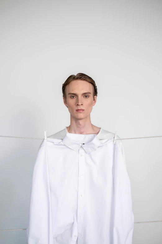 a man standing in front of a white wall, an album cover, inspired by Emanuel de Witte, wearing a white hospital gown, genderless, big collar, adam ondra