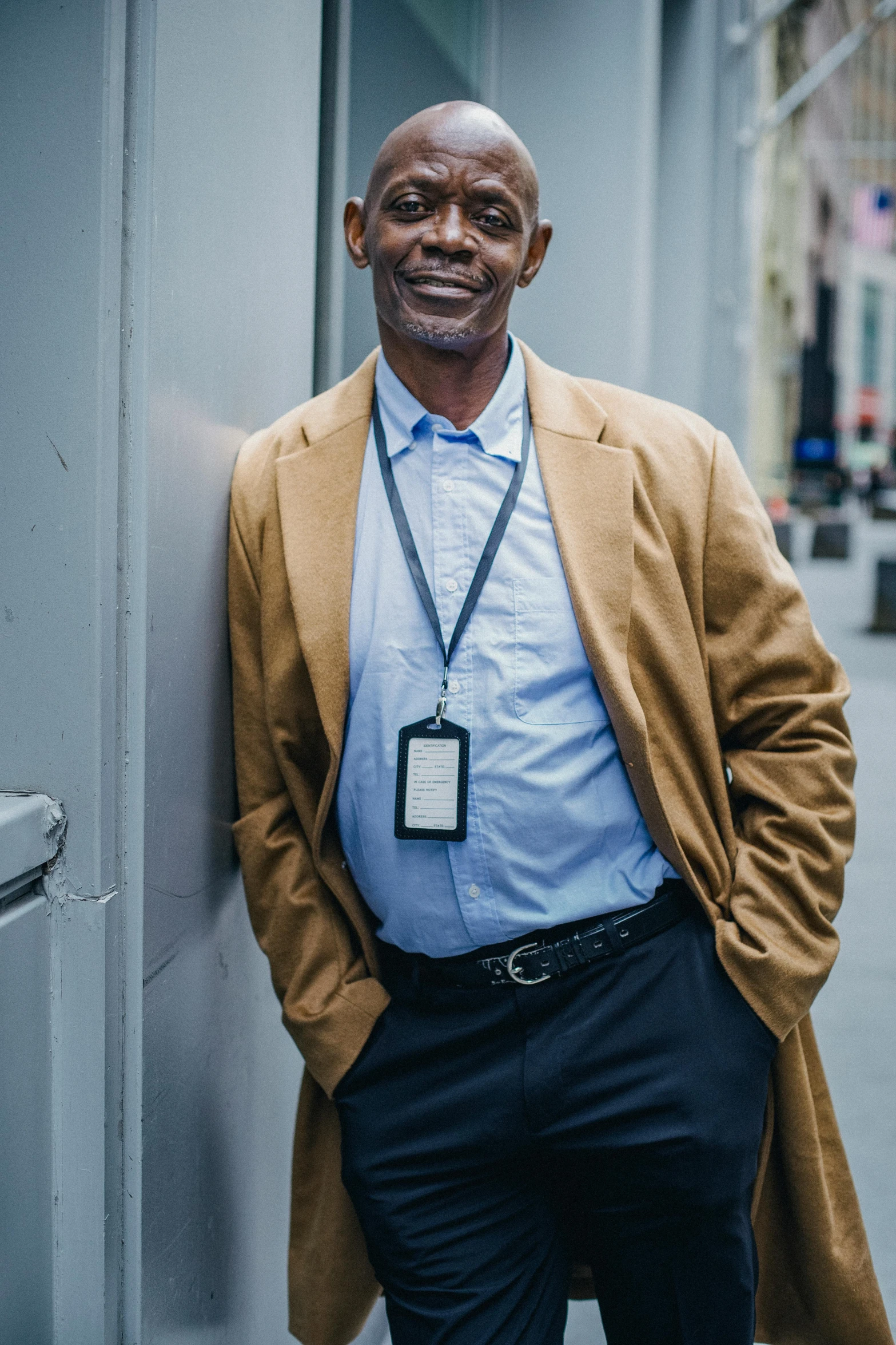 a man in a tan coat leaning against a wall, rodney matthew, standing in a city center, principal photography, profile image