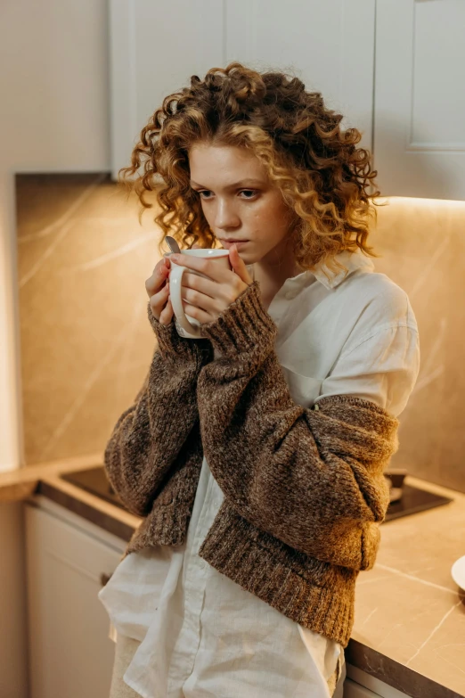 a woman standing in a kitchen holding a cup of coffee, a portrait, inspired by Elsa Bleda, trending on pexels, renaissance, short curly blonde haired girl, brown sweater, anna nikonova aka newmilky, cozy candlelight