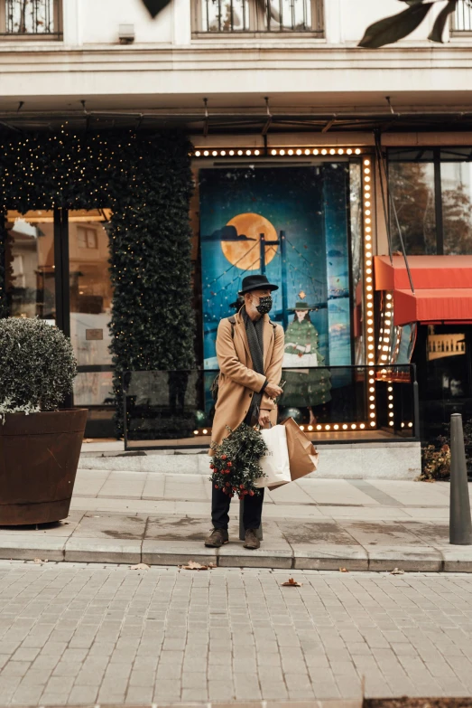 a man standing on a sidewalk in front of a building, by Julia Pishtar, pexels contest winner, wearing festive clothing, beige fedora, exiting store, wearing a hoodie and flowers