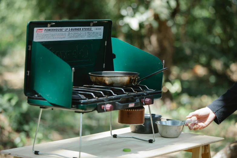 a close up of a person cooking food on a stove, by Emma Andijewska, private press, forest picnic, table in front with a cup, medium wide front shot, exterior shot