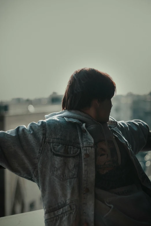 a man sitting on a ledge with his arms outstretched, an album cover, by Attila Meszlenyi, trending on pexels, happening, denim jacket, back light, red haired teen boy, showing her shoulder from back
