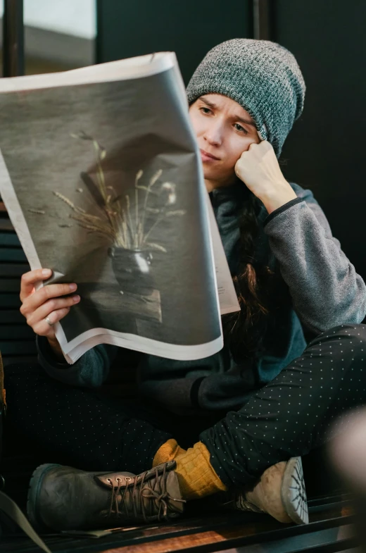 a woman sitting on the ground reading a newspaper, a picture, by Adam Marczyński, unsplash contest winner, wearing beanie, studio photo, bashful expression, print on magazine