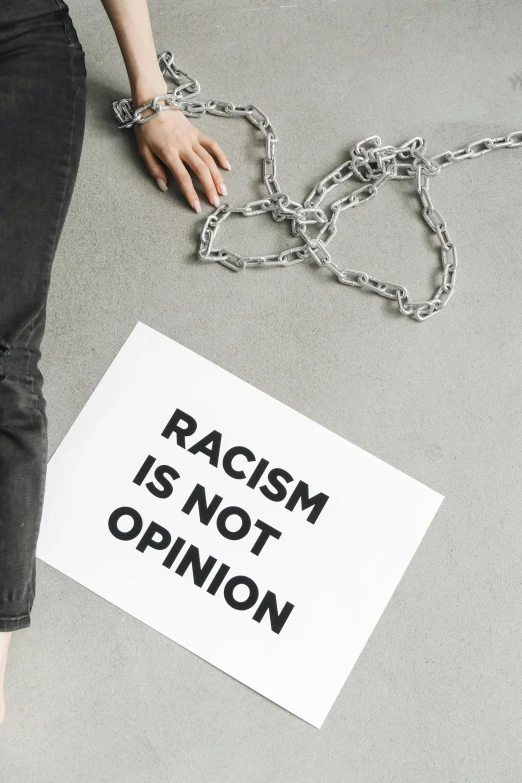 a woman is chained to a sign that says racism is not opinion, trending on unsplash, instagram post, splash image, jugendstill, etsy