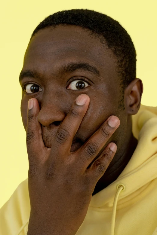 a man in a yellow hoodie covers his face with his hands, by Doug Ohlson, trending on pexels, hyperrealism, brown skin, looking surprised, hand on his cheek, lil uzi vert