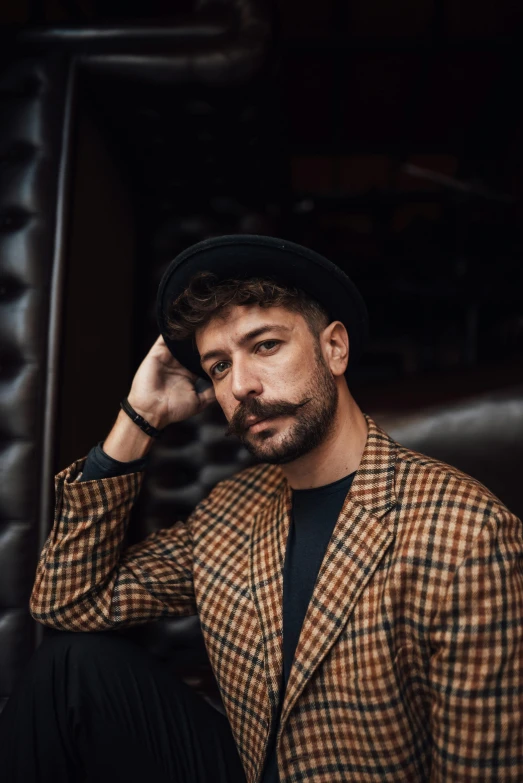 a man with a mustache sitting on a couch, an album cover, inspired by Germán Londoño, trending on pexels, caracter with brown hat, masculine pose, portrait of a young italian male, wearing a fancy jacket