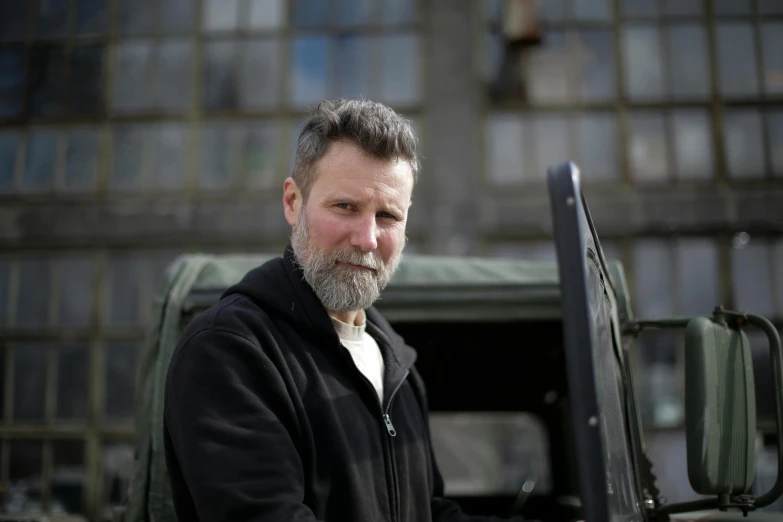 a man with a beard standing in front of a truck, a portrait, unsplash, aiden gillen, photographed for reuters, grey trimmed beard, in an urban setting