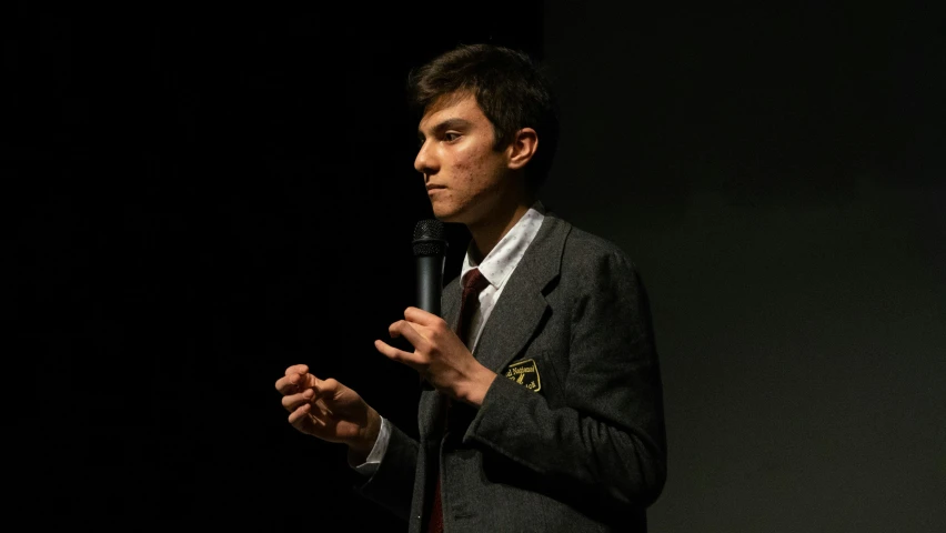 a man in a suit speaking into a microphone, liam brazier, thin young male, documentary photo, nathan fielder
